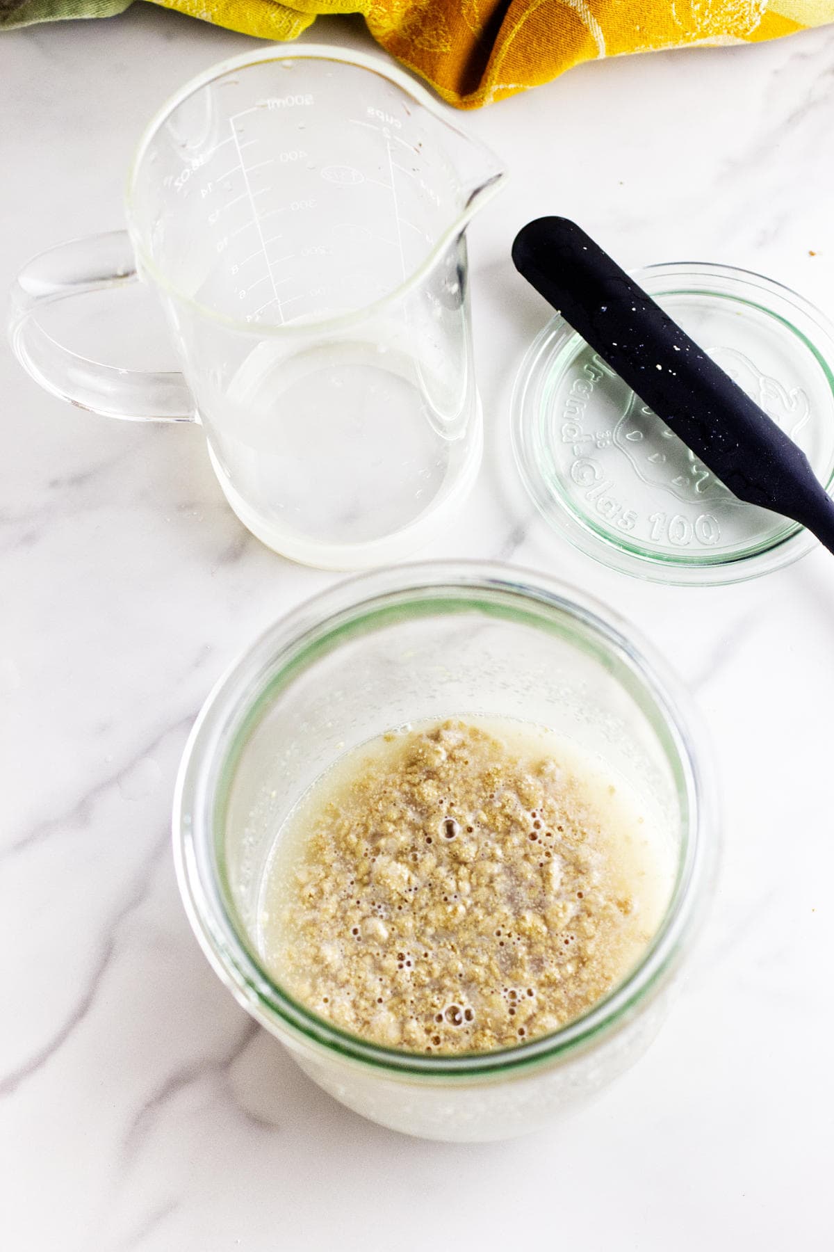 A glass jar filled with bubbling potato flake sourdough bread starter sits on a white surface next to an empty measuring cup, a black spatula, and a glass lid. A yellow cloth is partially visible in the corner.