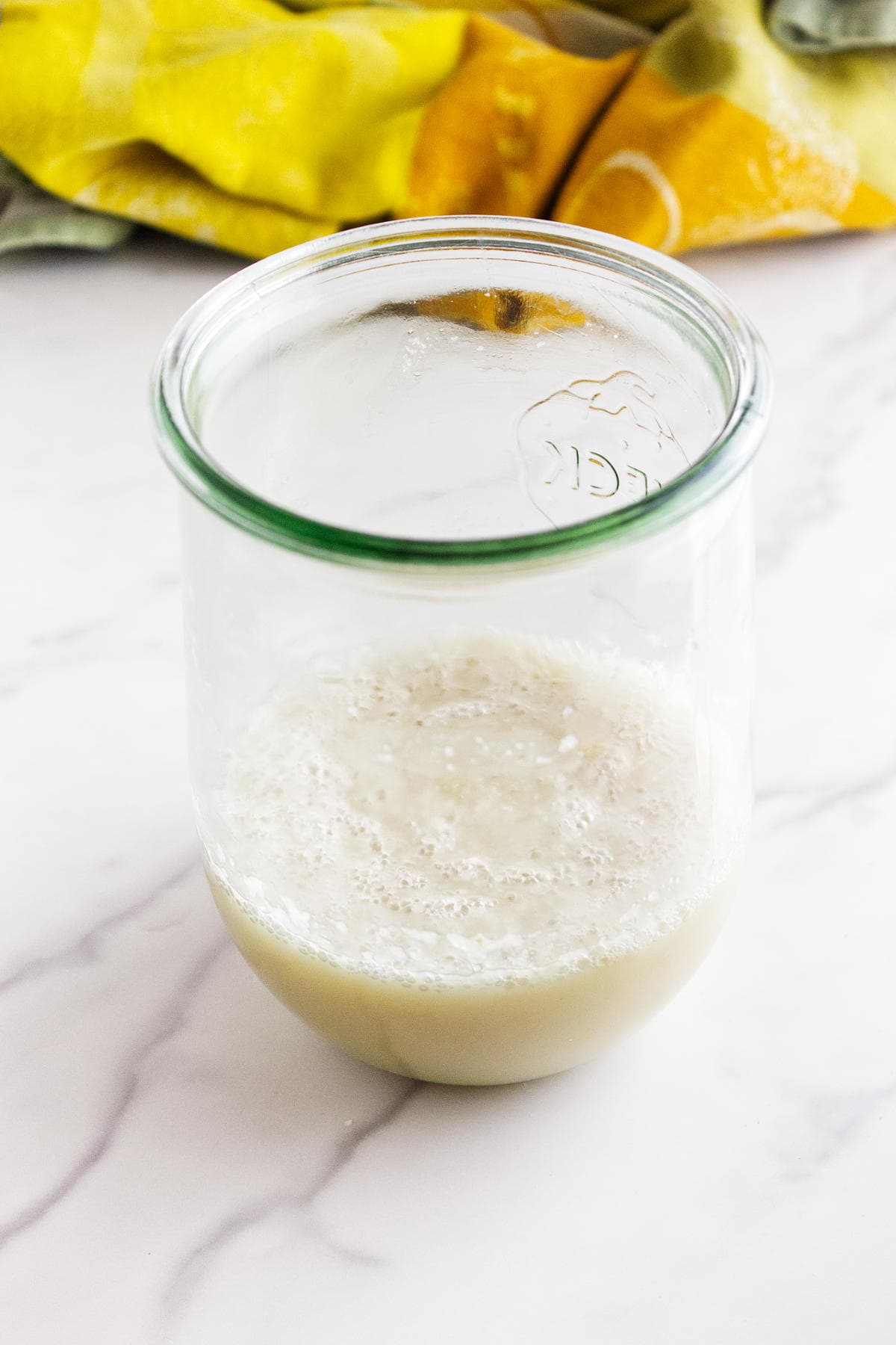 A glass jar with a foamy, light beige potato flake sourdough bread starter sits on a white marble surface. A yellow and green cloth is partially visible in the background.
