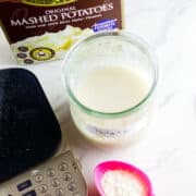 A jar labeled "Potato" filled with a white liquid sits on a counter next to a box of instant mashed potatoes, a digital kitchen scale, and a pink spoon holding potato flakes for making potato flake sourdough bread starter.
