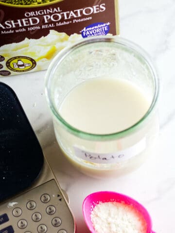 A jar labeled "Potato" filled with a white liquid sits on a counter next to a box of instant mashed potatoes, a digital kitchen scale, and a pink spoon holding potato flakes for making potato flake sourdough bread starter.