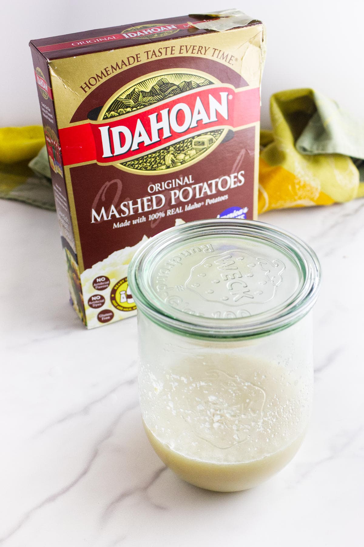 A box of Idahoan Original Mashed Potatoes sits behind a glass jar filled with prepared mashed potatoes, perfect for making potato flake sourdough bread starter, on a white surface, with a green and yellow cloth in the background.
