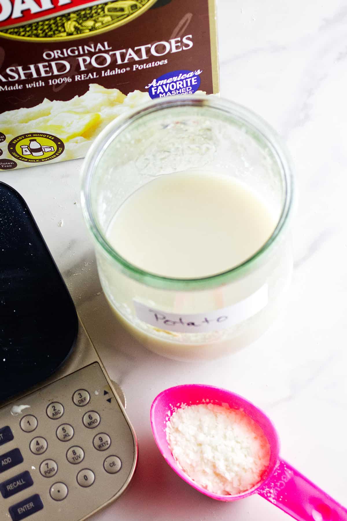 A jar labeled "Potato" contains a cloudy liquid, beside a pink spoon with white powder&mdash;possibly for a potato flake sourdough bread starter&mdash;a digital scale, and a box of instant mashed potatoes on a white surface.