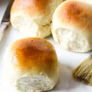Three golden brown potato flake sourdough rolls sit on a white surface next to a butter knife and pastry brush, showing their soft, fluffy texture and shiny, slightly glossy tops.