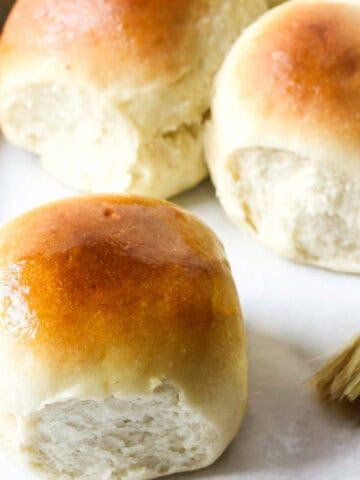 Three golden brown potato flake sourdough rolls sit on a white surface next to a butter knife and pastry brush, showing their soft, fluffy texture and shiny, slightly glossy tops.