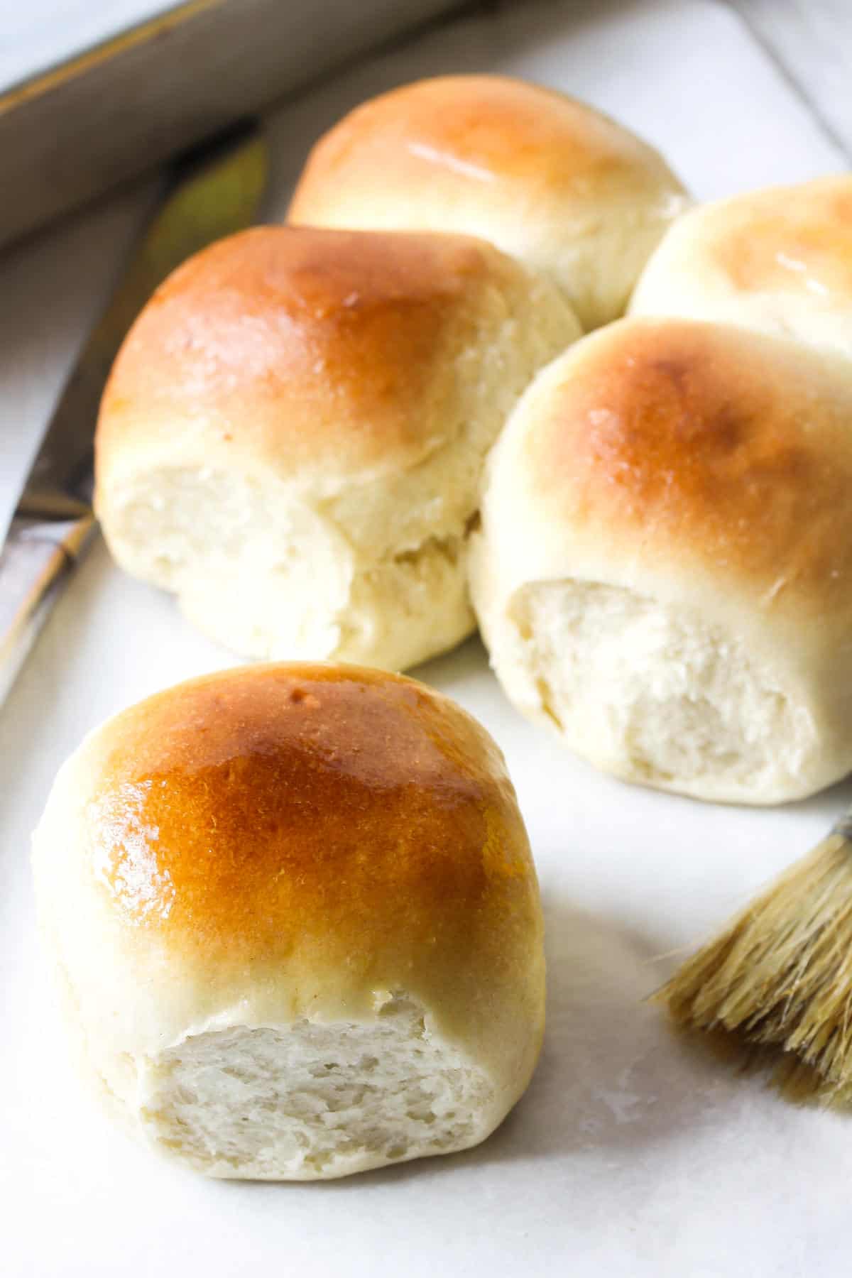 Four golden-brown, fluffy potato flake sourdough rolls are arranged on a light surface next to a butter knife and pastry brush. The rolls have a shiny, slightly glazed top.