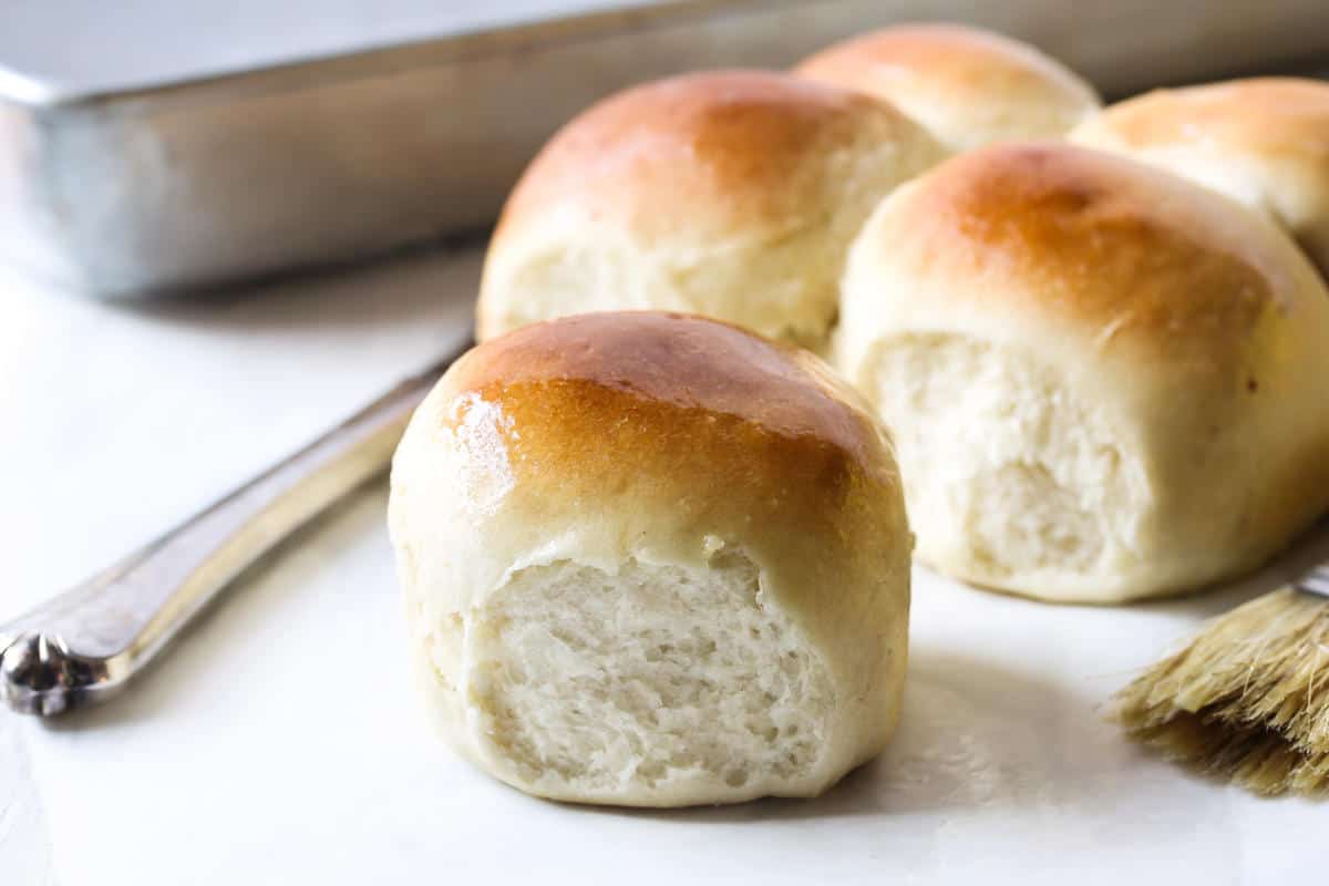 Freshly baked golden brown potato flake sourdough rolls on a white surface, with a baking pan, fork, and pastry brush nearby. The rolls have a soft and fluffy texture.