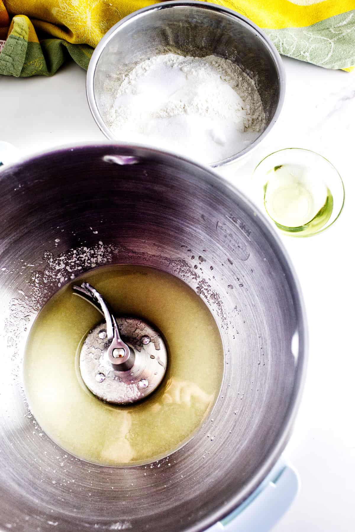 A stand mixer bowl with liquid and a dough hook, ready for potato flake sourdough bread, sits beside a metal bowl of flour and a small glass bowl of yeast on a white countertop with a green and yellow towel in the background.