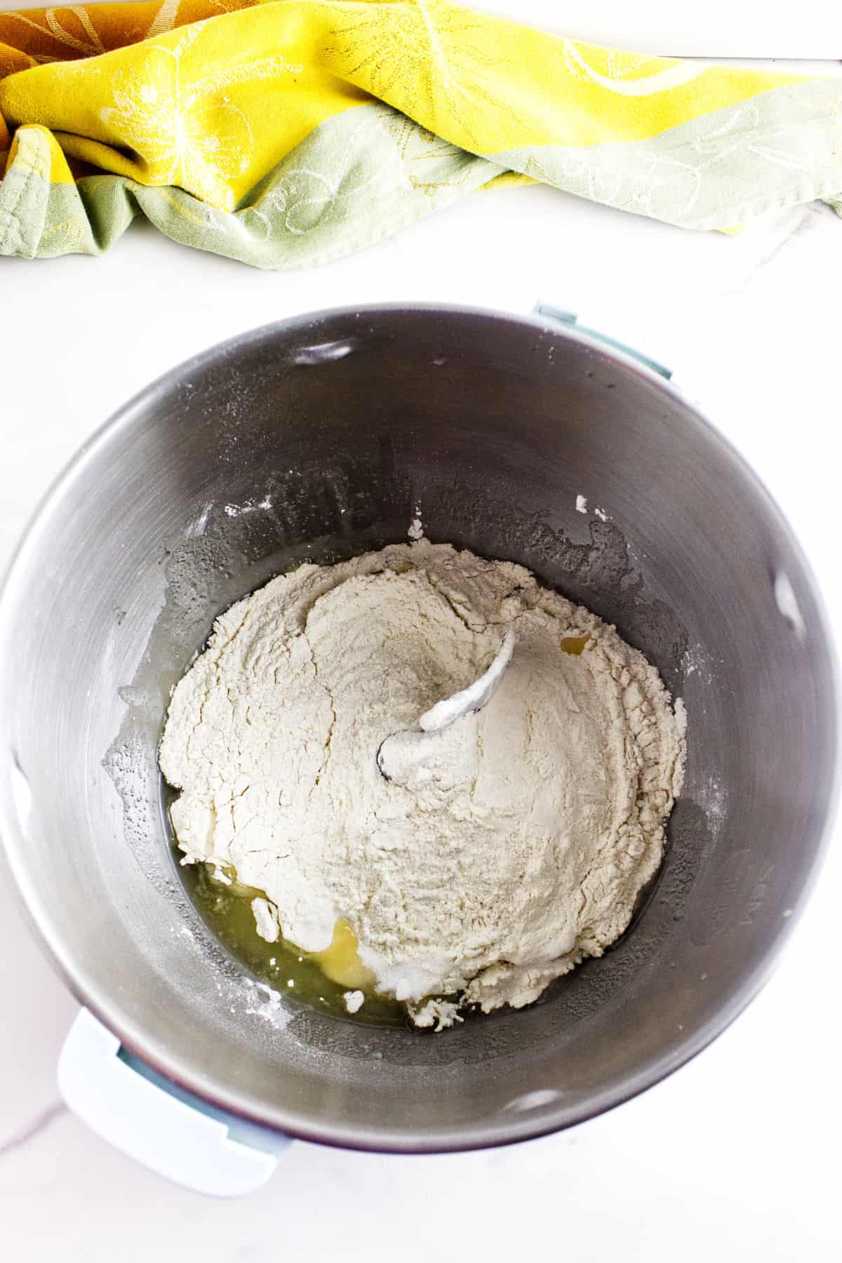 A mixing bowl with flour, water, and a small amount of oil for potato flake sourdough bread sits on a white surface with a yellow and green cloth in the background, ready to be mixed.