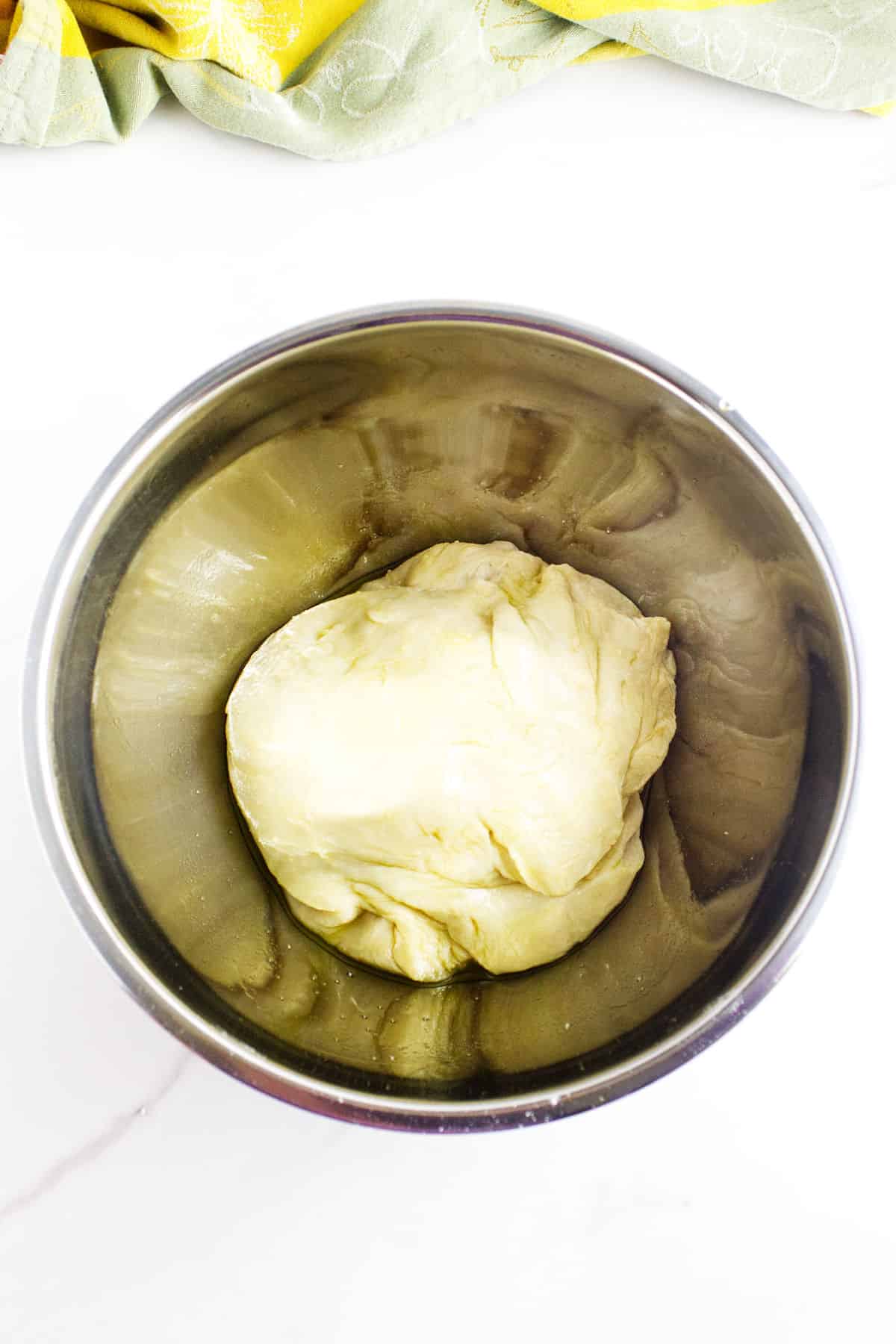 A stainless steel bowl containing a ball of potato flake sourdough bread dough rests on a white surface, with a yellow and green cloth partially visible at the top of the image.
