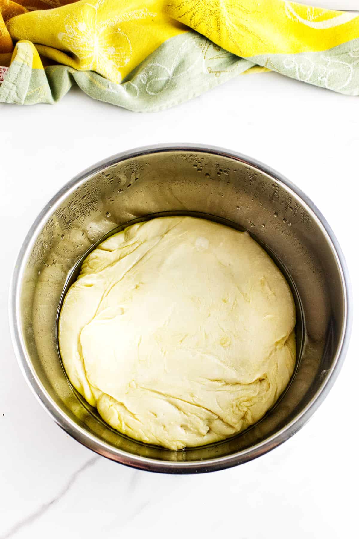 A stainless steel bowl filled with risen potato flake sourdough bread dough, coated in oil, sits on a white surface with a colorful yellow and green cloth nearby.