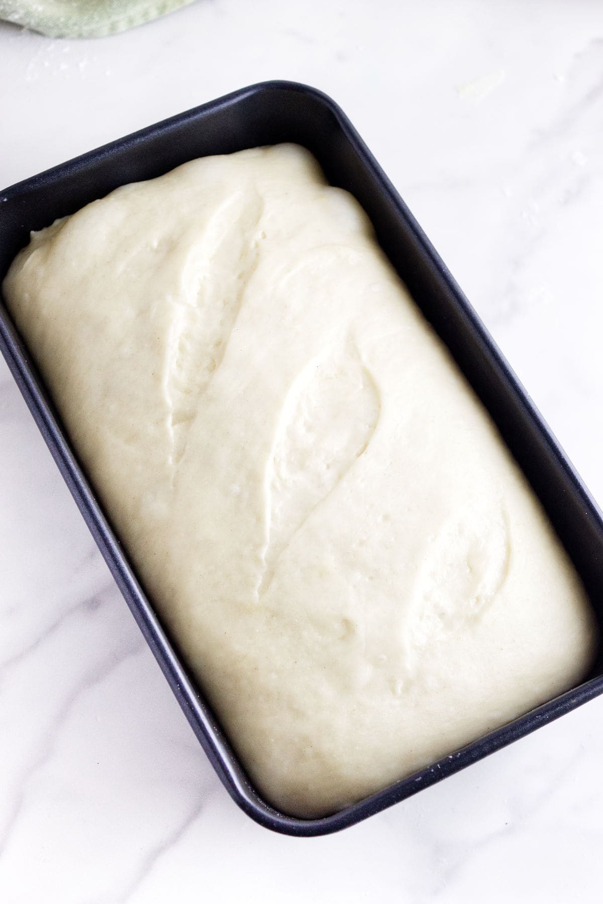 A loaf pan filled with uncooked, risen potato flake sourdough bread dough rests on a white marble surface. The dough features three leaf-shaped slashes on top.