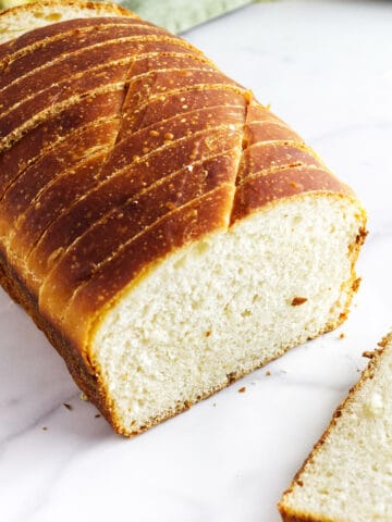 A loaf of sliced potato flake sourdough bread with a golden-brown crust sits on a white surface, with one slice lying next to it.