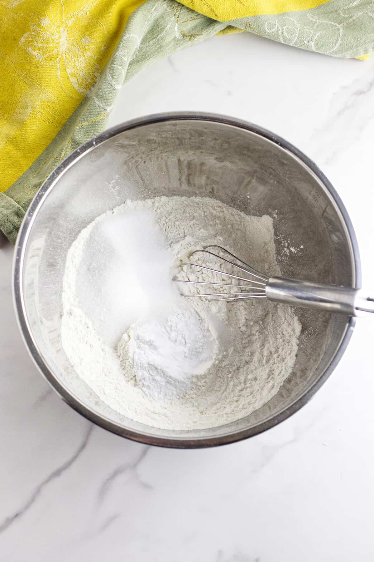 A metal mixing bowl with flour, sugar, and baking powder being whisked together for rhubarb scones, placed on a marble countertop with a yellow and green kitchen towel nearby.