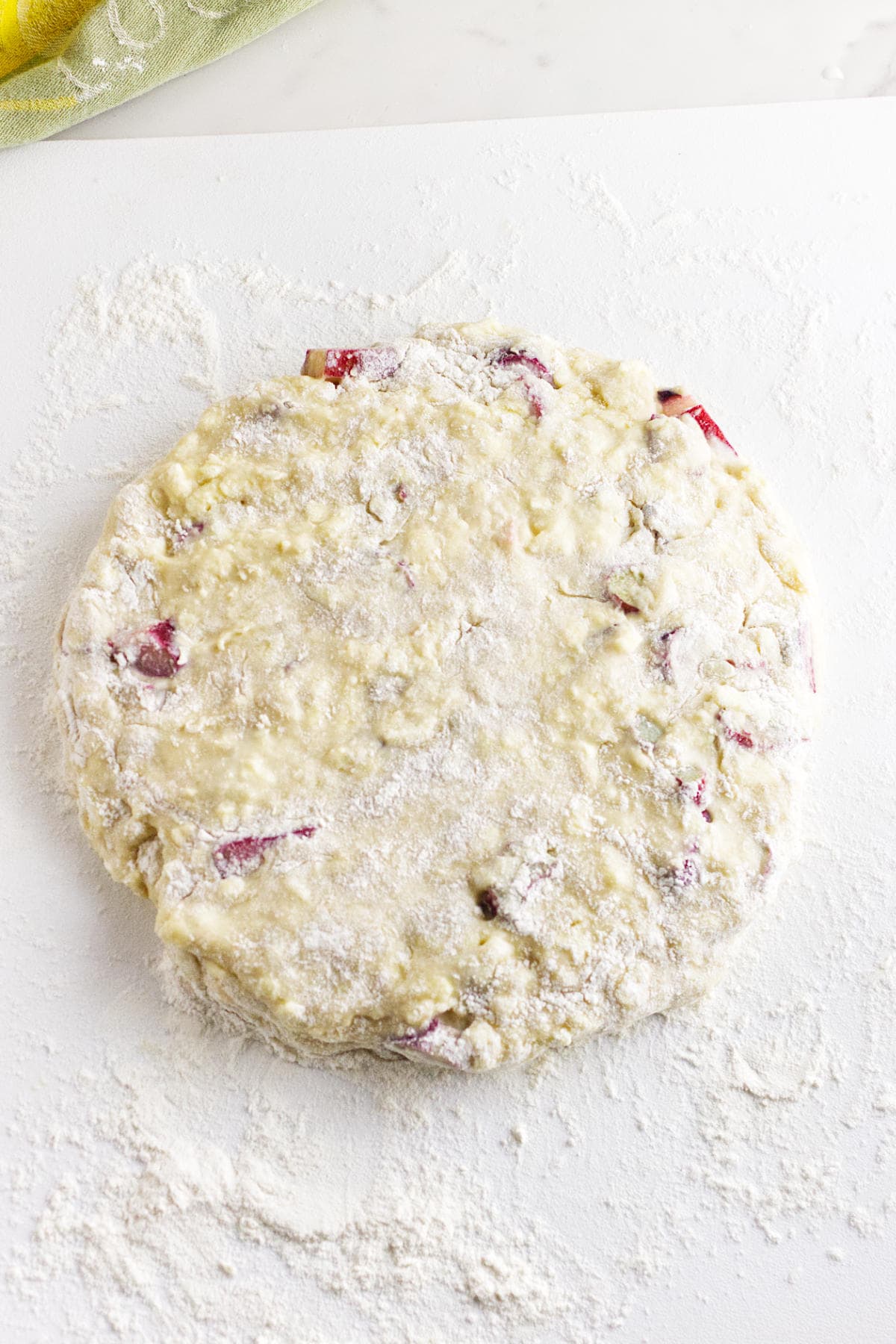 A round disk of unbaked dough with visible pieces of red fruit, perfect for rhubarb scones, rests on a floured white surface, ready to be shaped or cut. Flour is scattered around the dough.
