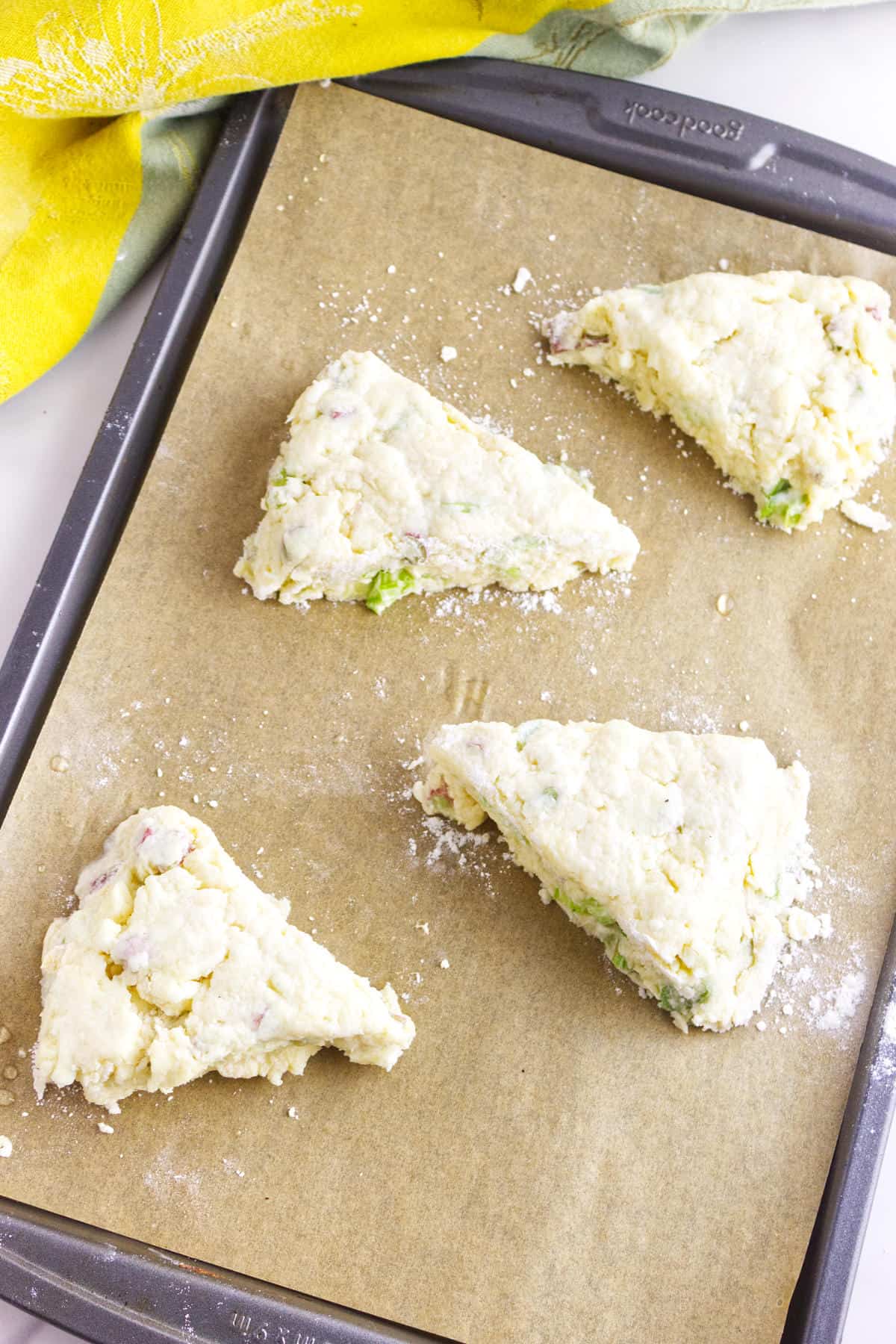 Four unbaked, triangular rhubarb scones with visible chunks of avocado and bacon sit on a parchment-lined baking sheet, ready to be baked. A yellow and green cloth is partially visible in the corner.
