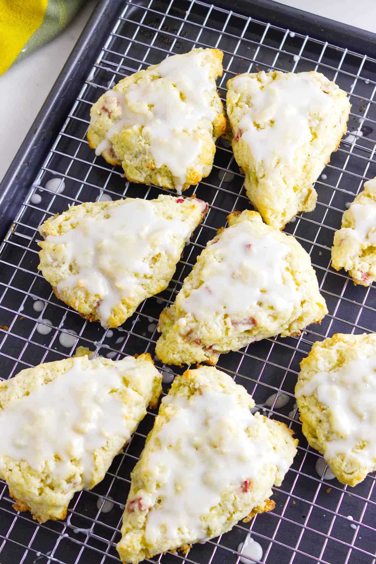Eight triangular rhubarb scones with white glaze are arranged on a wire cooling rack placed over a baking sheet. The scones appear golden brown, with small bits of rhubarb showing through the dough.