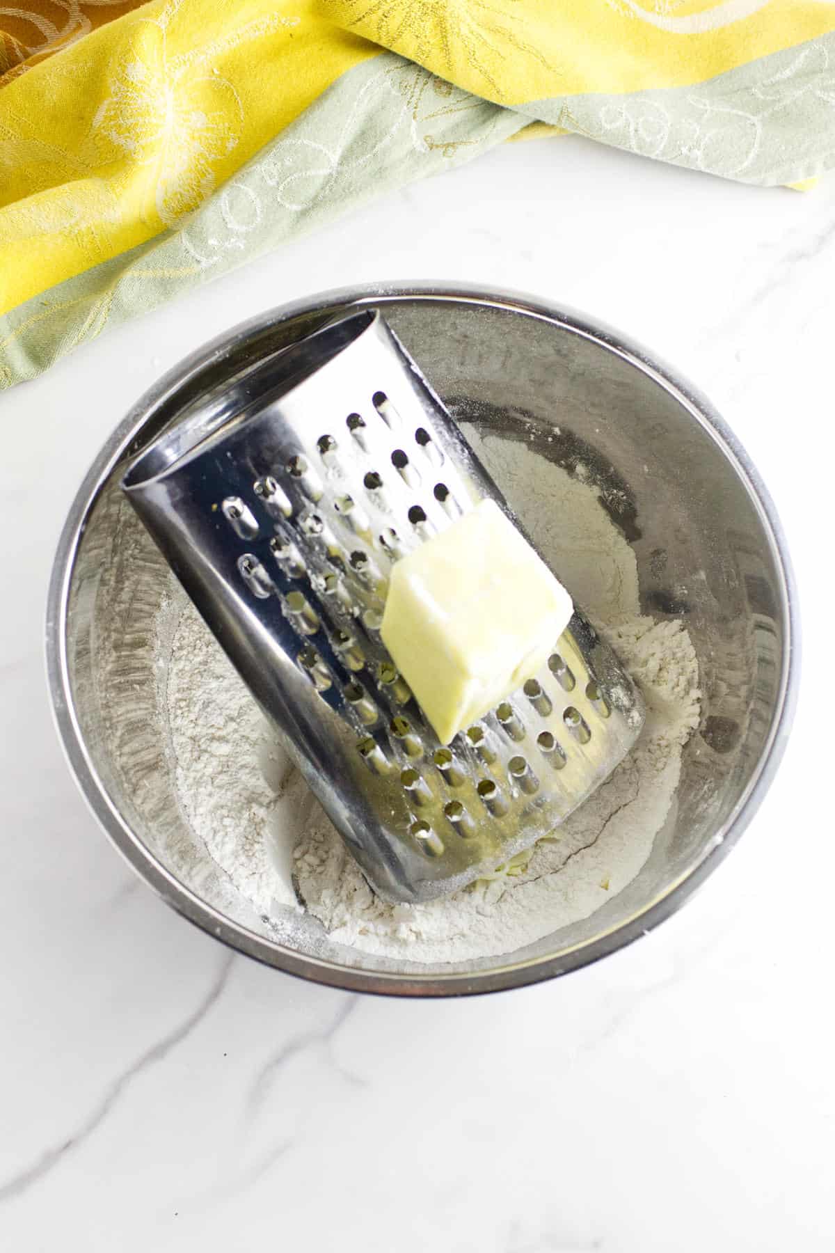 A metal box grater with a stick of butter being grated over a mixing bowl filled with flour for rhubarb scones, set on a white marble surface with a yellow and green cloth nearby.