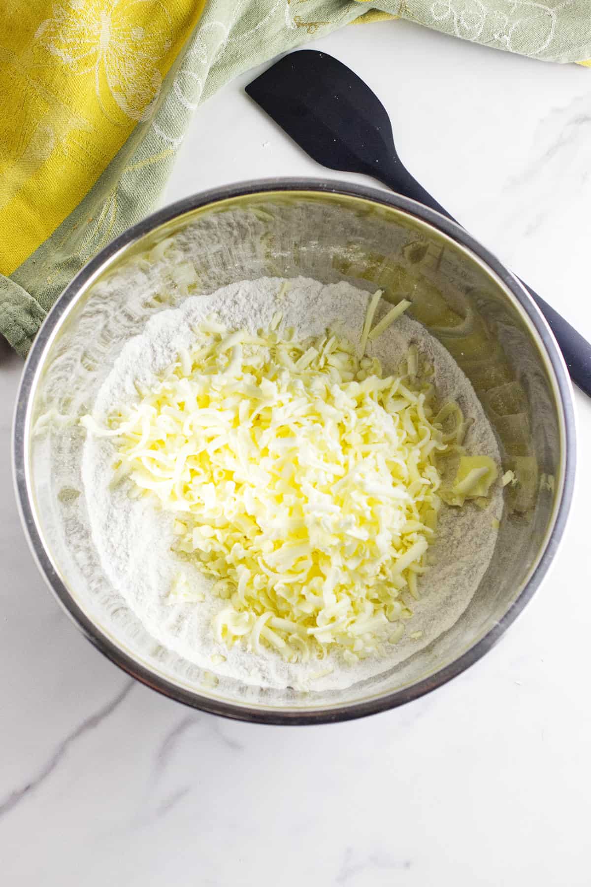 A metal mixing bowl with flour and shredded butter for rhubarb scones sits on a marble countertop next to a black spatula and a green kitchen towel.
