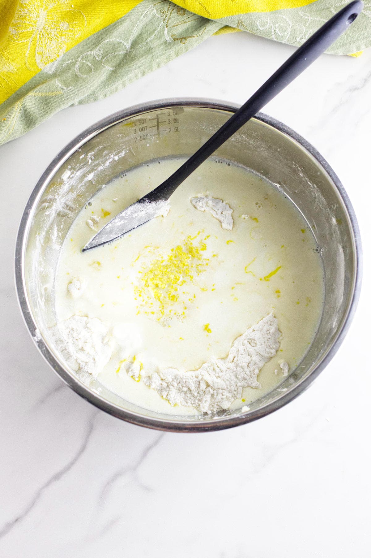 A metal mixing bowl contains flour, milk, lemon zest, and other ingredients for rhubarb scones being mixed with a black spatula on a white marble countertop. A yellow patterned cloth is partially visible in the background.