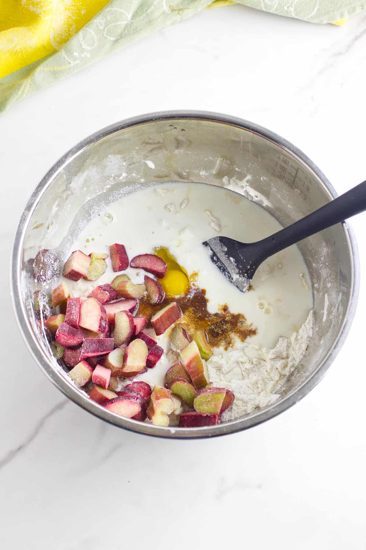 A metal mixing bowl with chopped rhubarb, an egg, liquid ingredients, flour, and spices being mixed for rhubarb scones with a black spatula on a white marble surface.