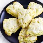 Six glazed rhubarb scones are arranged on a black plate. The scones are golden with a light icing on top and have a slightly rough, homemade texture.