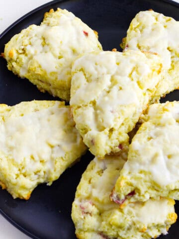 Six glazed rhubarb scones are arranged on a black plate. The scones are golden with a light icing on top and have a slightly rough, homemade texture.