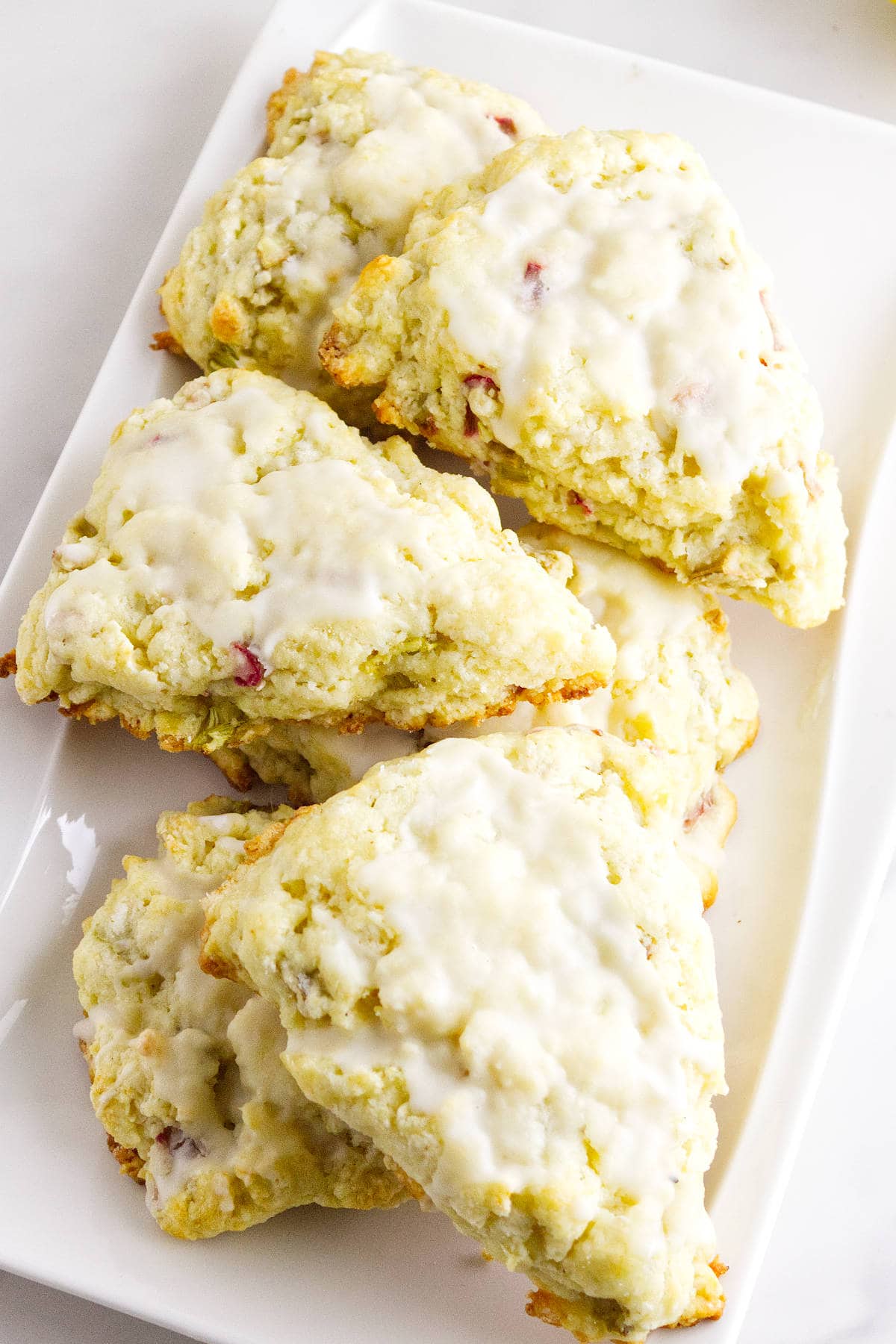 A white rectangular plate holds several triangular rhubarb scones topped with a light white glaze. The scones have a crumbly texture, and small bits of fruit can be seen within the dough.