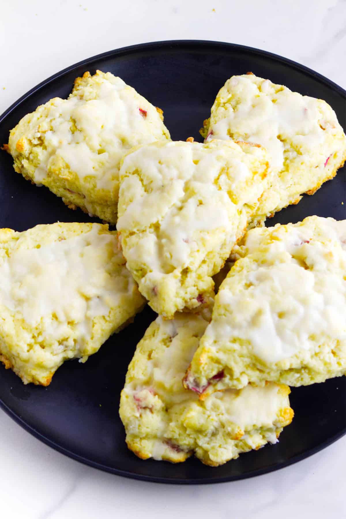 Six glazed rhubarb scones are arranged on a black plate. The scones are golden with a light icing on top and have a slightly rough, homemade texture.