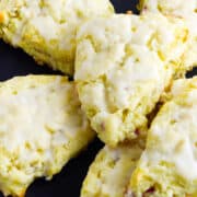 Six glazed rhubarb scones are arranged on a black plate. The scones are golden with a light icing on top and have a slightly rough, homemade texture.