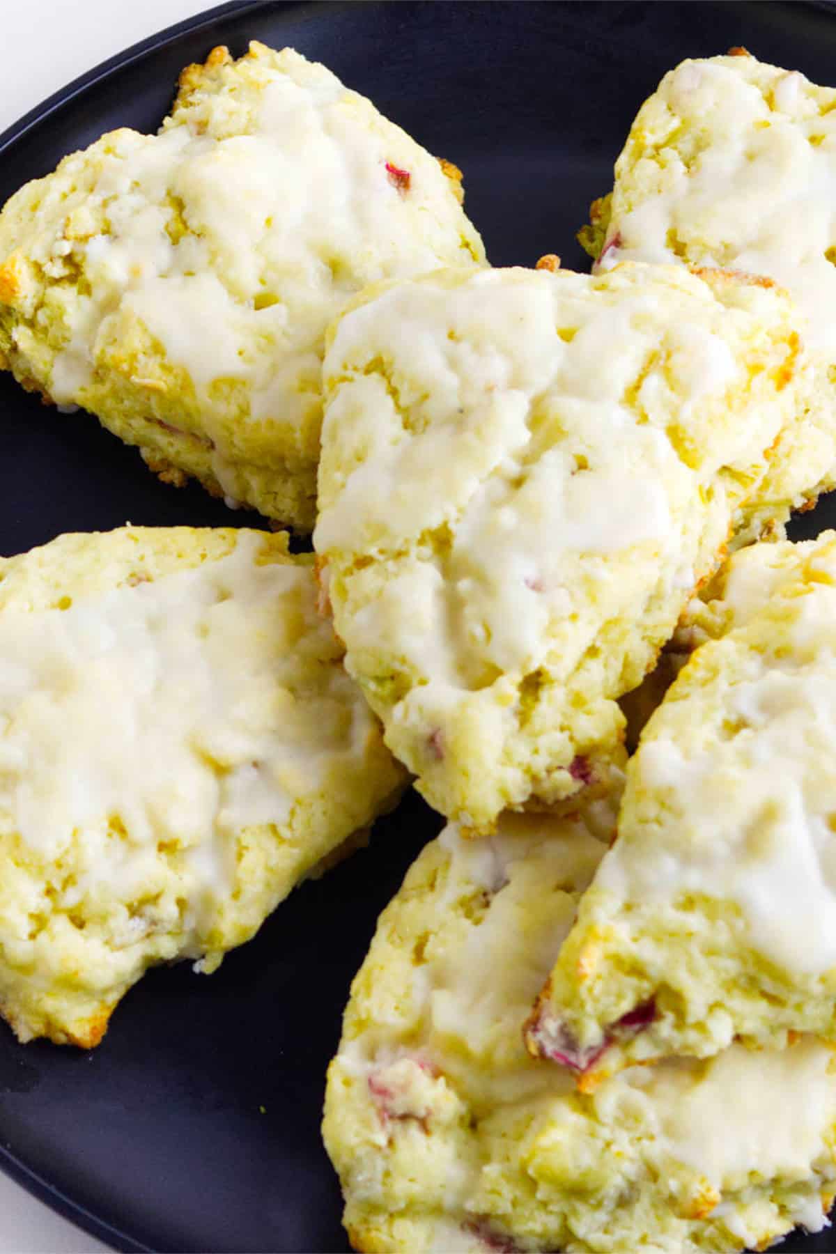 Six glazed rhubarb scones are arranged on a black plate. The scones are golden with a light icing on top and have a slightly rough, homemade texture.