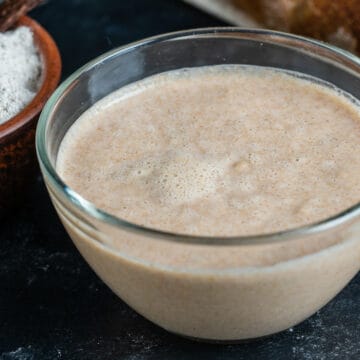 A clear glass bowl filled with a bubbly, beige sourdough starter sits on a dark surface beside a bowl of flour.
