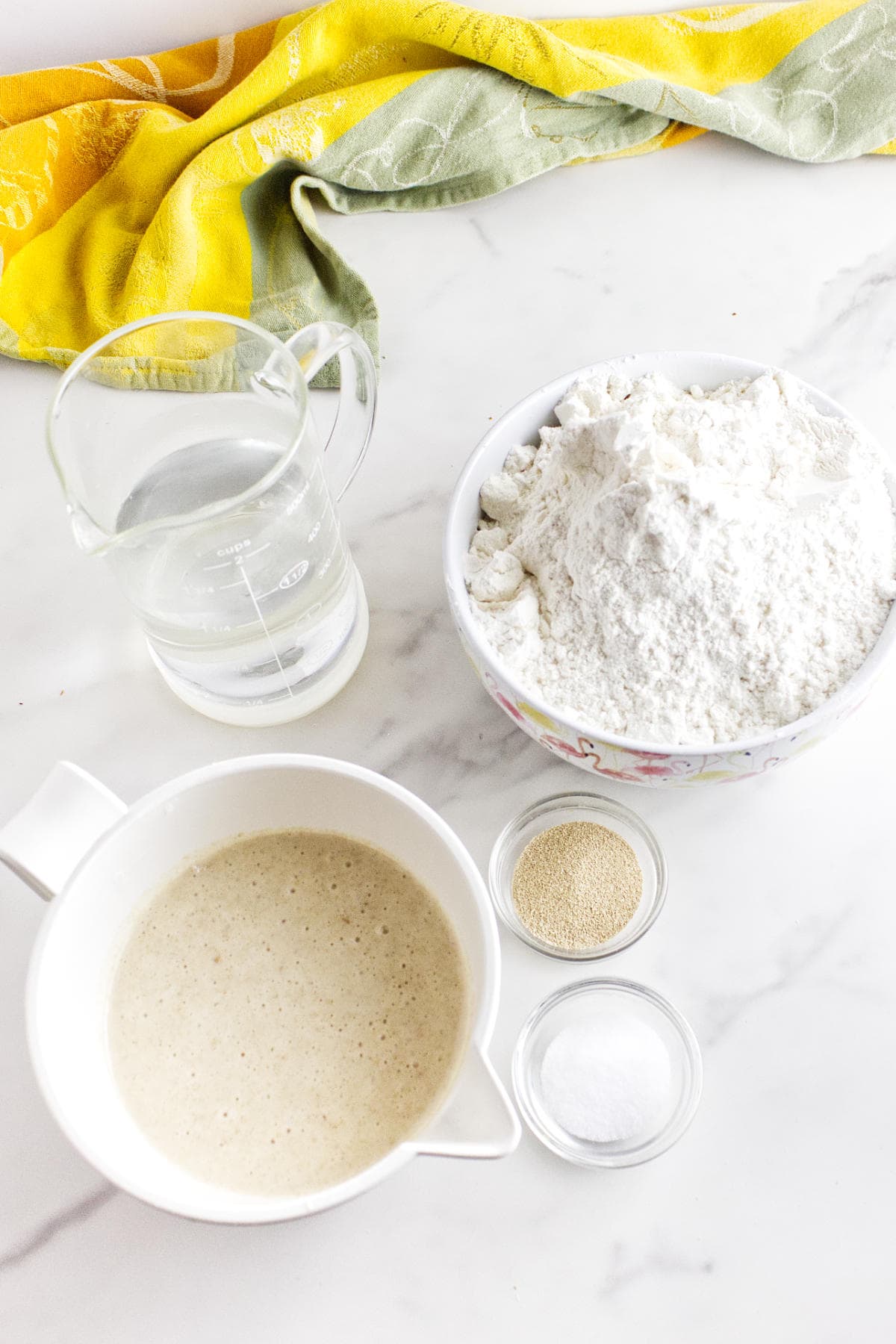 On a white surface, bowls with flour and bubbly white sourdough discard starter sit beside dishes of yeast and salt, a clear measuring cup with water, and a yellow-green kitchen towel—perfect for making soft sandwich bread.