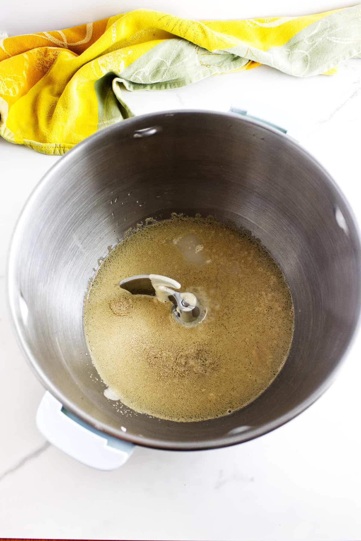 A mixing bowl with yeast and water being mixed for white sourdough discard sandwich bread, placed on a white countertop with a yellow and green kitchen towel in the background.