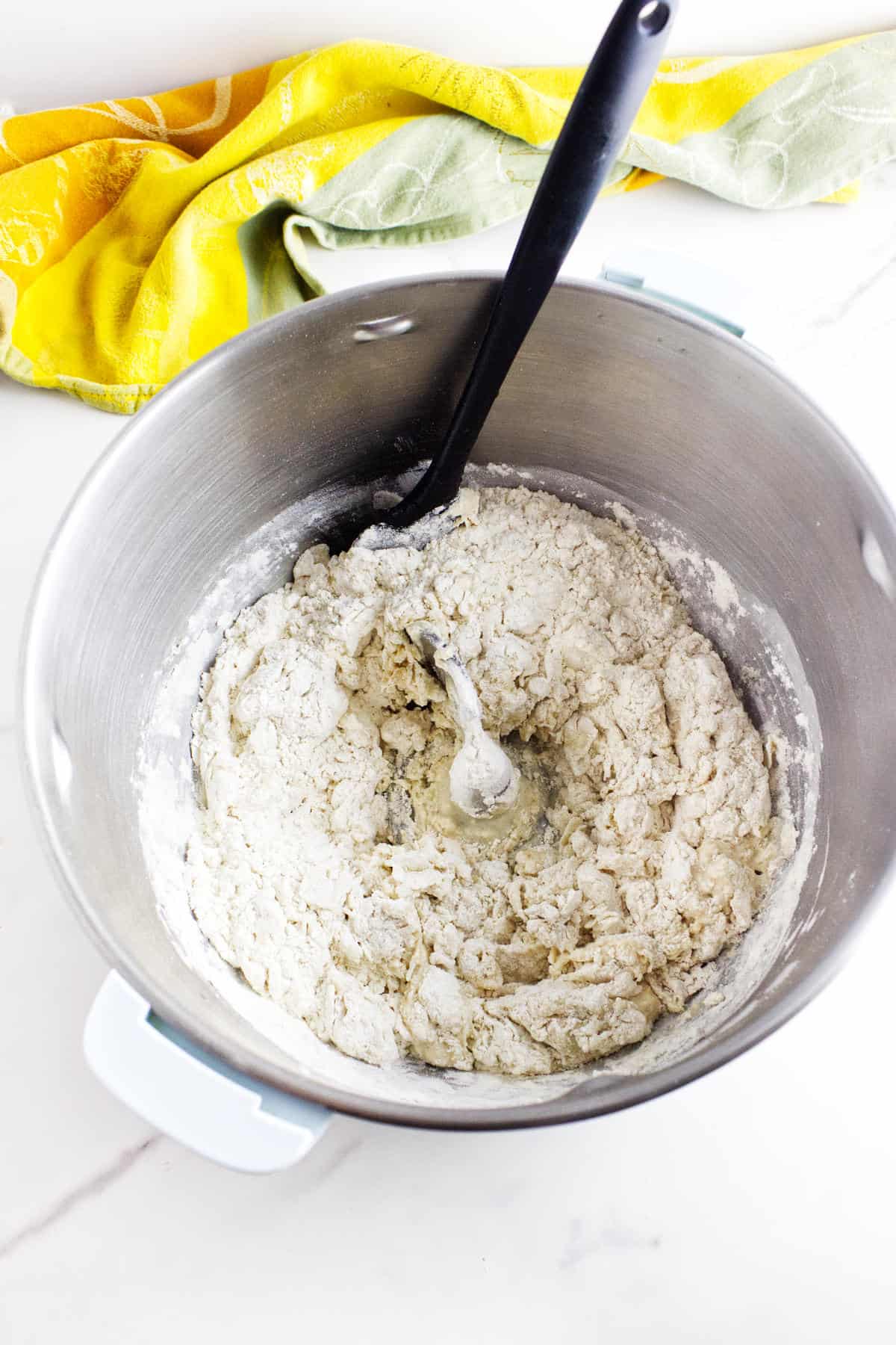A metal mixing bowl with partially mixed white sourdough discard sandwich bread dough and a black spoon inside, set on a white surface with a yellow and green kitchen towel nearby.