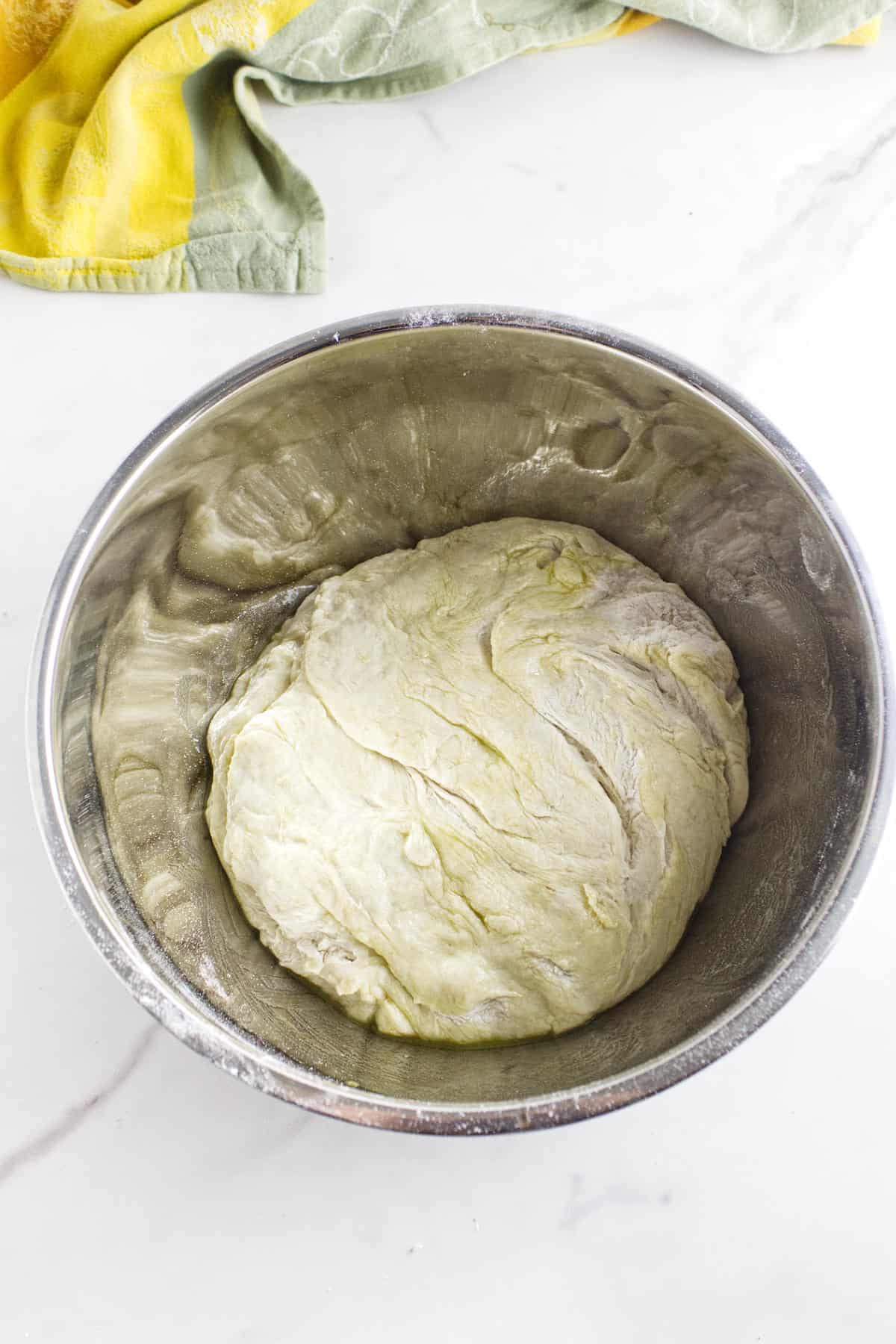 A ball of white sourdough discard sandwich bread dough rests in a large metal bowl on a white countertop, with a yellow and green kitchen towel partially visible in the corner.