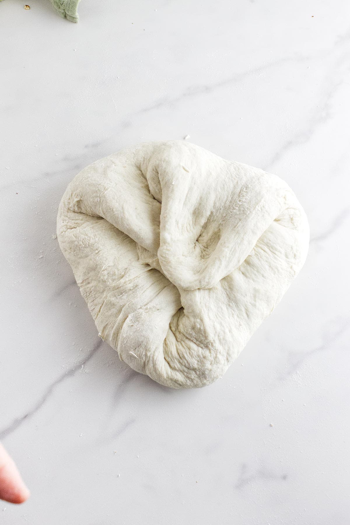 A ball of raw white sourdough discard sandwich bread dough with folded edges rests on a white marble surface. A fingertip is visible in the lower left corner of the image.