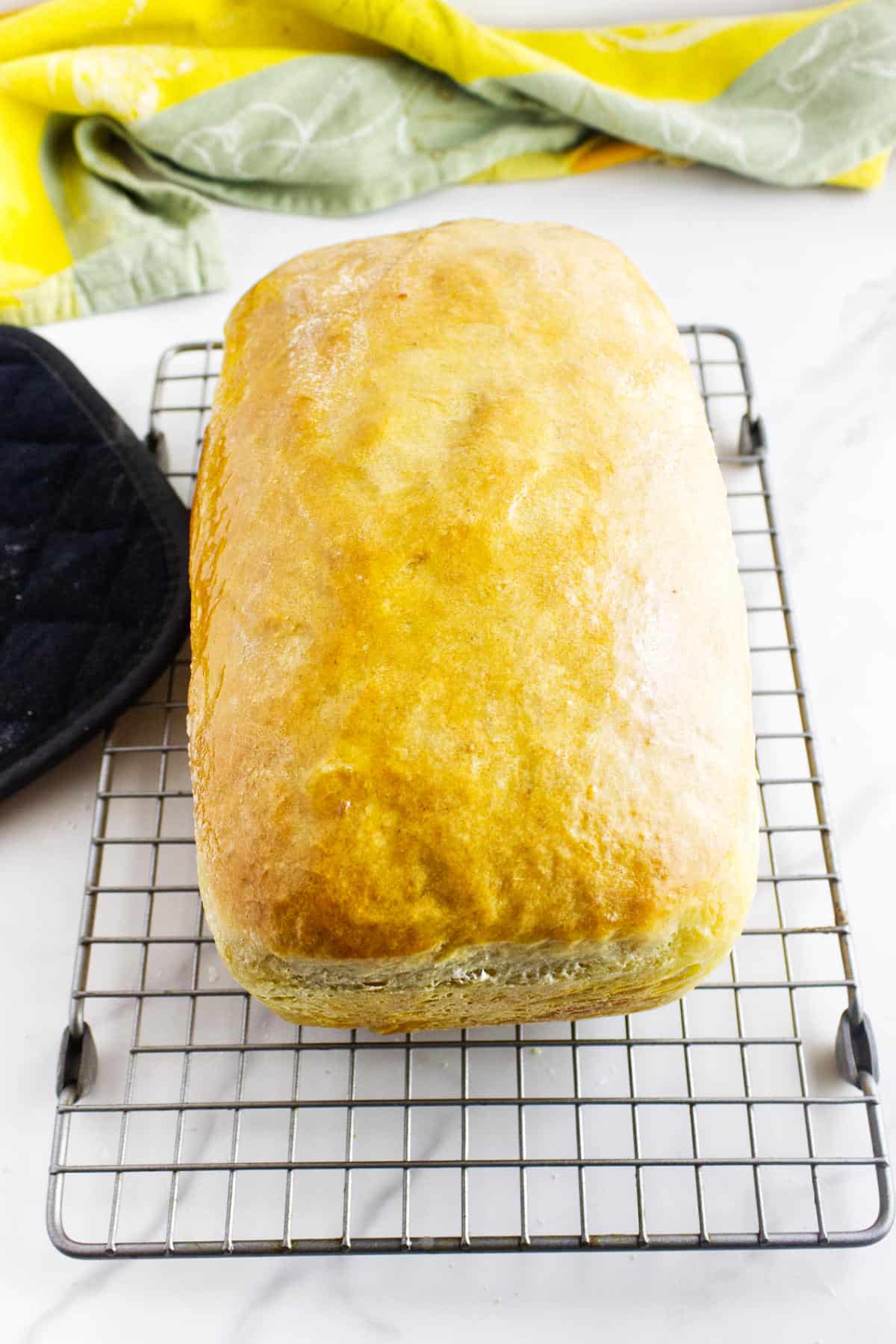 A golden brown loaf of white sourdough discard sandwich bread cools on a wire rack, with a black oven mitt to the left and a green and yellow kitchen towel in the background.