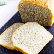 A loaf of homemade white sourdough discard sandwich bread with a golden crust sits on a dark cutting board, two thick slices cut and laid in front.