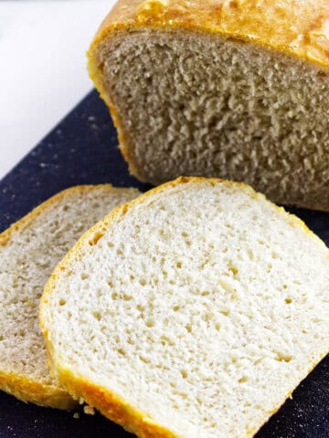 A loaf of homemade white sourdough discard sandwich bread with a golden crust sits on a dark cutting board, two thick slices cut and laid in front.