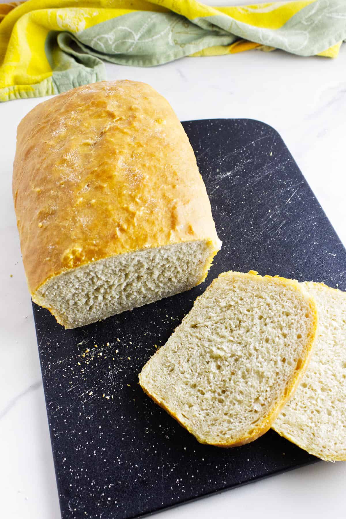 A loaf of homemade white sourdough discard sandwich bread sits on a black cutting board with two slices cut in front. A green and yellow kitchen towel is in the background on a white countertop.