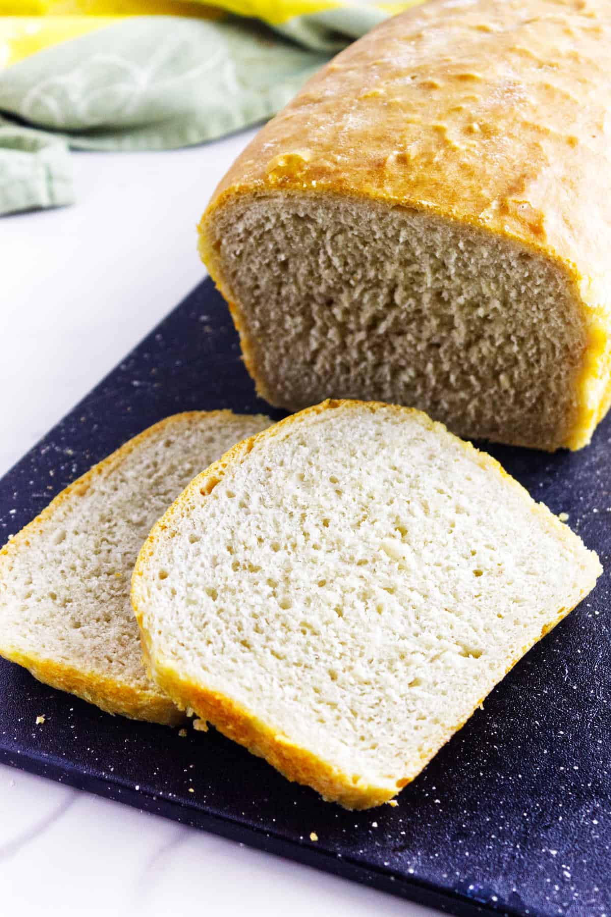 A loaf of homemade white sourdough discard sandwich bread sits on a dark cutting board with two slices cut in front, showing a soft, airy texture and golden crust. A green cloth is visible in the background.