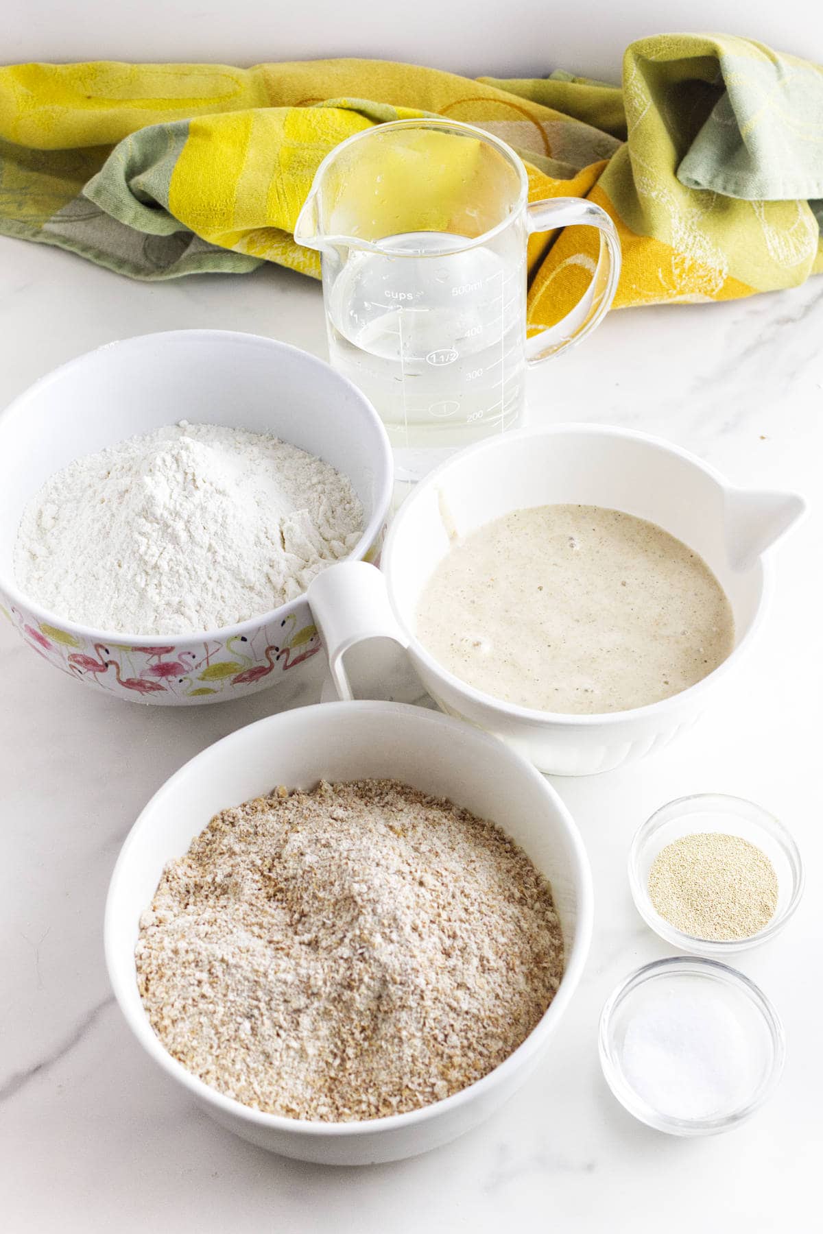 Bowls of ingredients for bread: white flour, wheat flour, yeast mixture, and small bowls of yeast and salt, plus water—perfect for making whole wheat sourdough discard sandwich bread—set on a white surface with a yellow-green cloth.