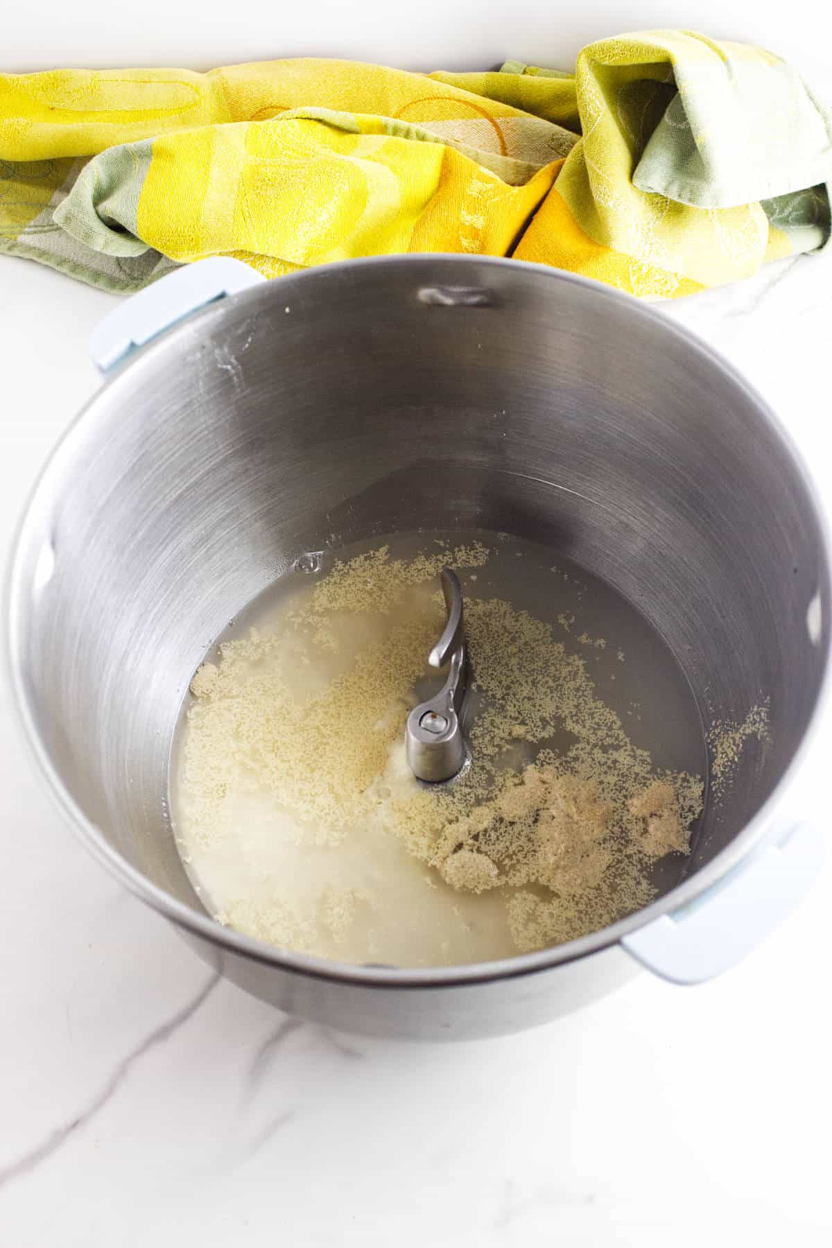 A stand mixer bowl with water and yeast, ready for whole wheat sourdough discard sandwich bread, with a dough hook attached. A yellow and green kitchen towel sits in the background on a white countertop.