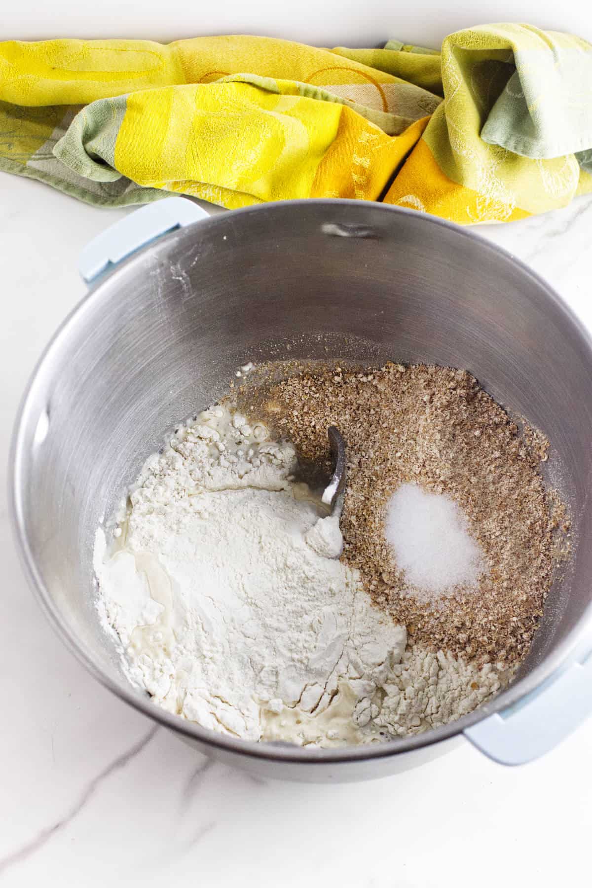 A large metal mixing bowl containing flour, ground flaxseed, and salt for whole wheat sourdough discard sandwich bread sits on a white countertop, with a yellow and green kitchen towel in the background.