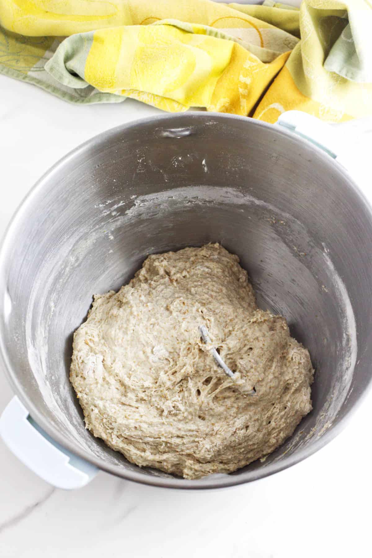 A metal mixing bowl filled with sticky, partially mixed whole wheat sourdough discard sandwich bread dough sits on a white countertop, with a colorful yellow and green kitchen towel in the background.