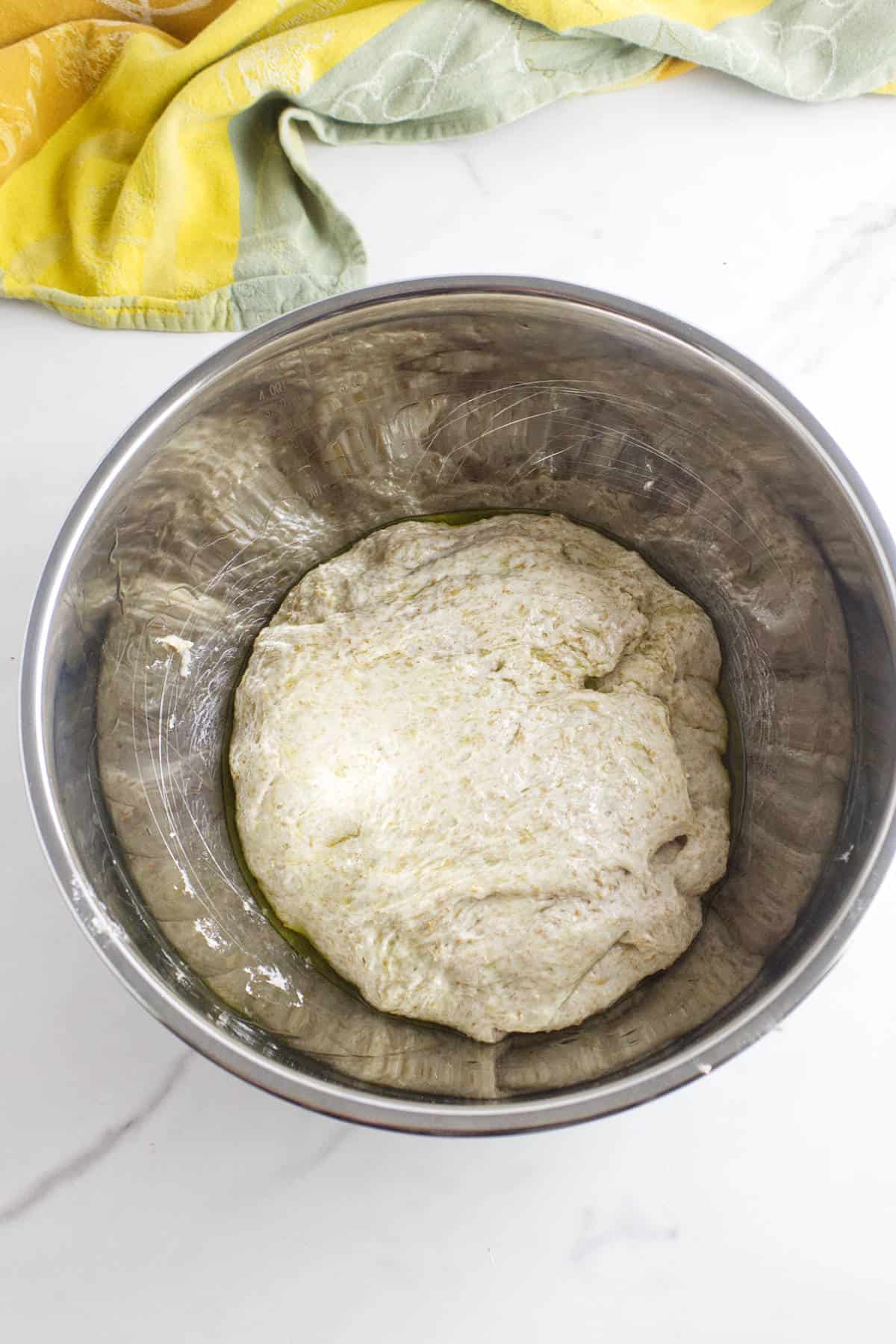 A stainless steel bowl containing risen whole wheat sourdough discard sandwich bread dough sits on a white marble surface, with a yellow and green kitchen towel partially visible at the top of the image.