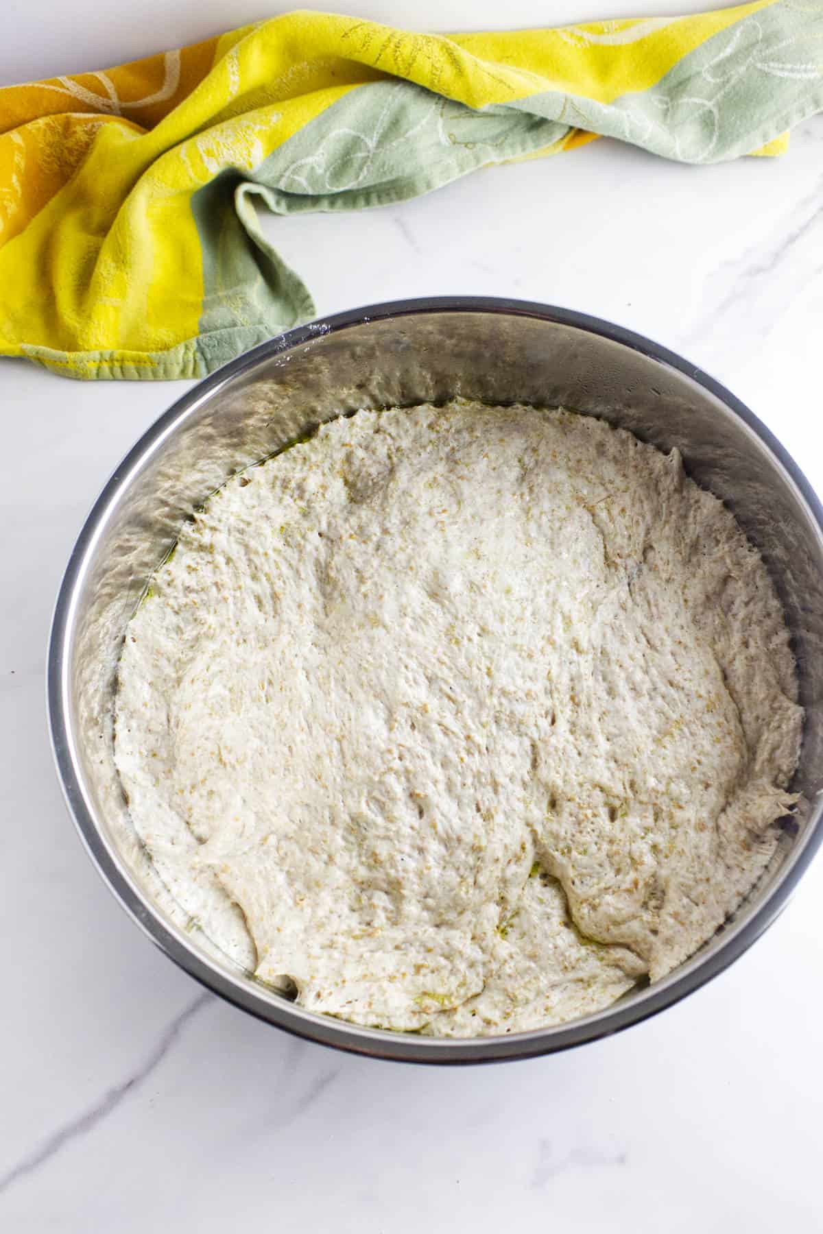 A large metal bowl filled with risen whole wheat sourdough discard sandwich bread dough sits on a white marble surface, with a yellow and green kitchen towel in the background.