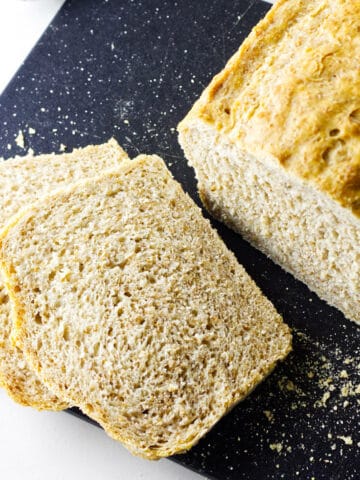A loaf of whole wheat sourdough discard sandwich bread rests on a black cutting board, two slices cut and lying beside it. Bread crumbs are scattered across the surface.