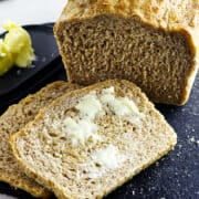 A loaf of whole wheat sourdough discard sandwich bread sits on a black cutting board with two slices in front, one spread with melting butter. More butter rests on a small dish nearby.