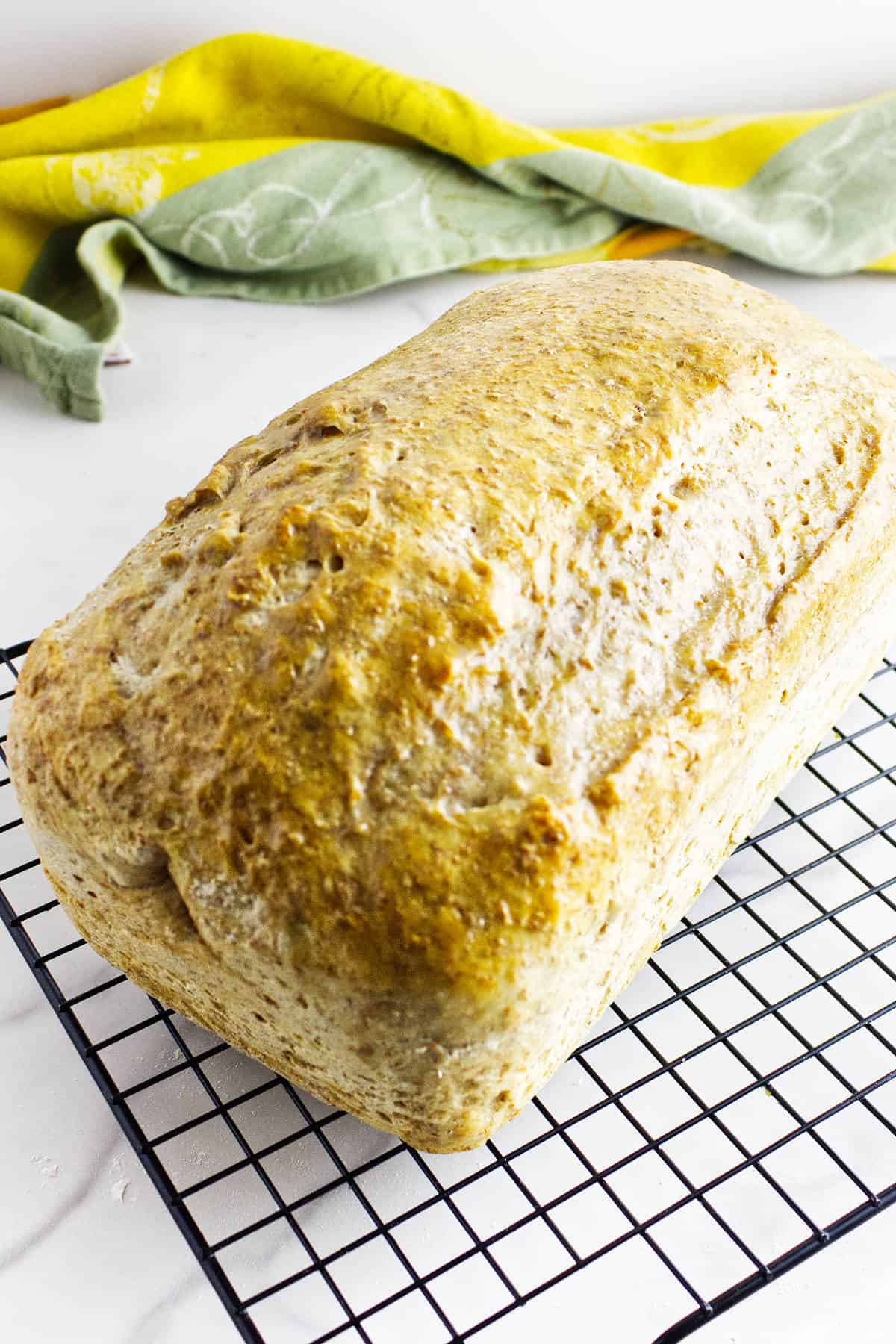 A golden-brown loaf of whole wheat sourdough discard sandwich bread rests on a black wire cooling rack, with a yellow and green kitchen towel in the background on a white surface.